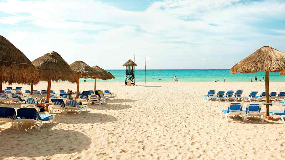 Hiekkaranta, jossa aurinkotuoleja ja varjoja sekä meri taustalla. (Tekoälyn luoma teksti)