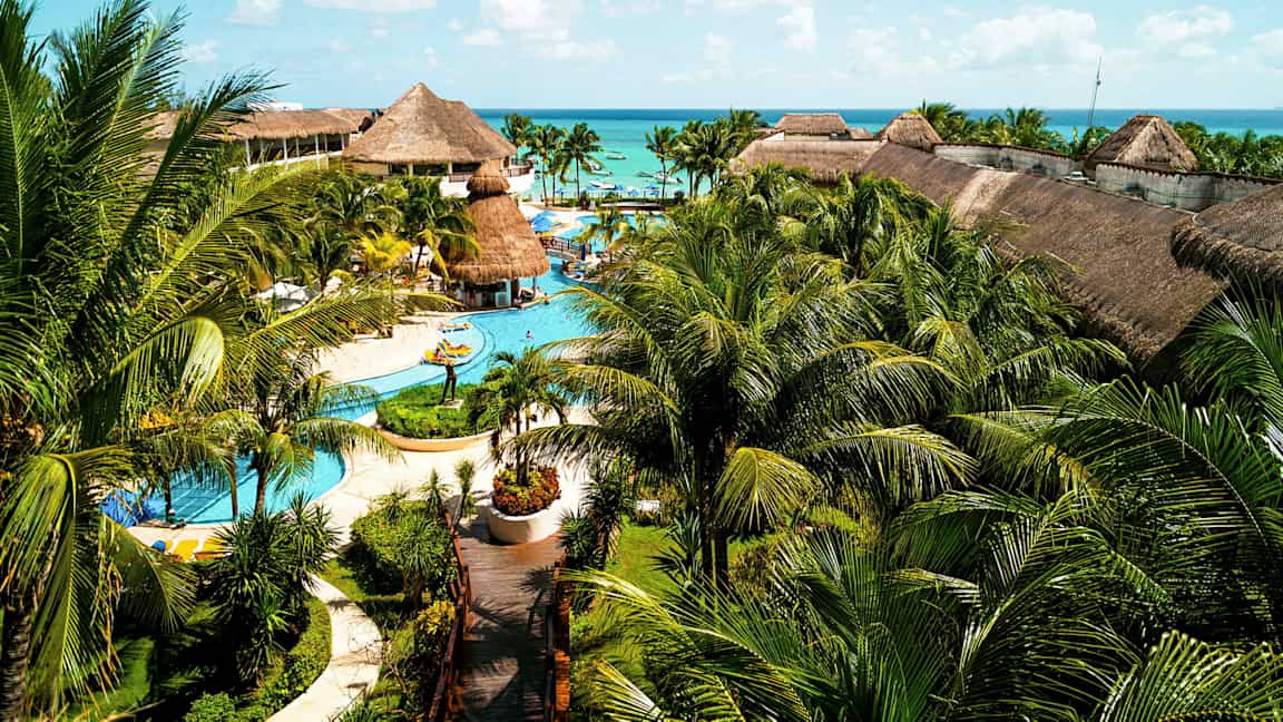 Suuri lomakeskus, jossa on uima-altaita, palmuja ja olkikattoisia rakennuksia, sijaitsee lähellä merta sinisen taivaan alla. (Tekoälyn luoma teksti)