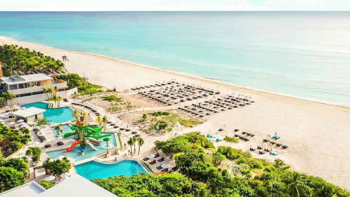 Kuva hiekkarannasta meren äärellä ja sinisen veden ympäröimänä. Rannalla on aurinkotuoleja ja aurinkovarjoja. Etualalla näkyy uima-allas vesiliukumäkien kera sekä ympäröivä vihreä kasvillisuus. (Tekoälyn luoma teksti)