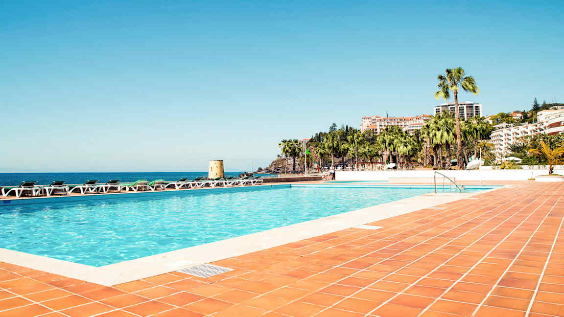 Uima-allas ympäröitynä aurinkotuoleilla, näkymä merelle ja rakennuksia palmujen keskellä. (Tekoälyn luoma teksti)