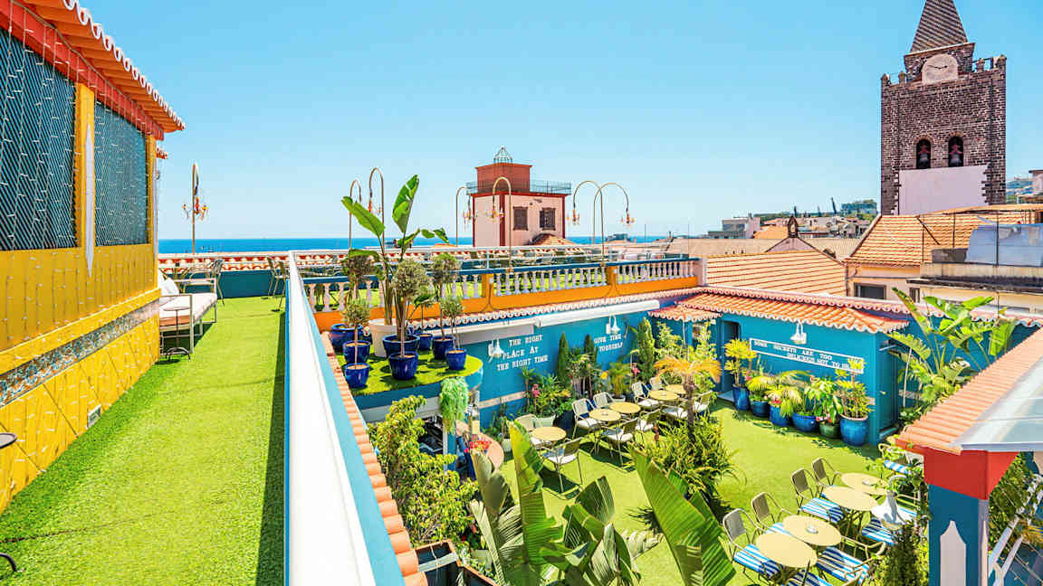 Kattoterassi, jossa on vihreitä kasveja, istumapaikkoja ja värikkäitä rakennuksia taustalla. (Tekoälyn luoma teksti)