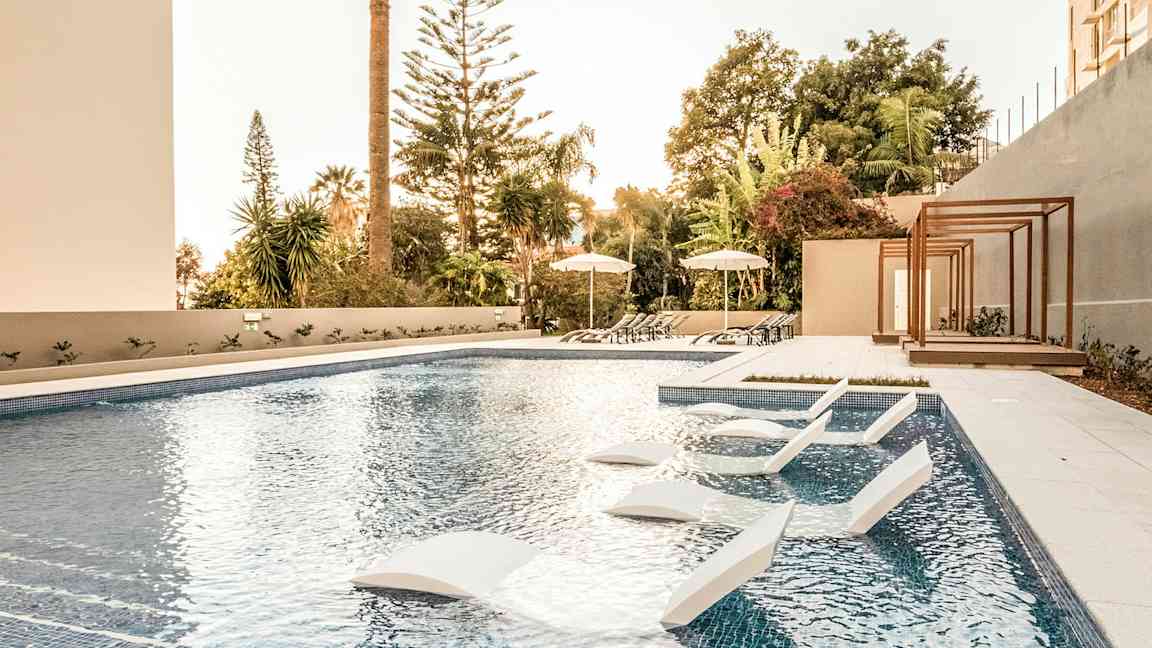 Uima-allas ulkona, jossa aurinkotuoleja ja varjoja, ympäröivät puut ja kasvillisuus. (Tekoälyn luoma teksti)