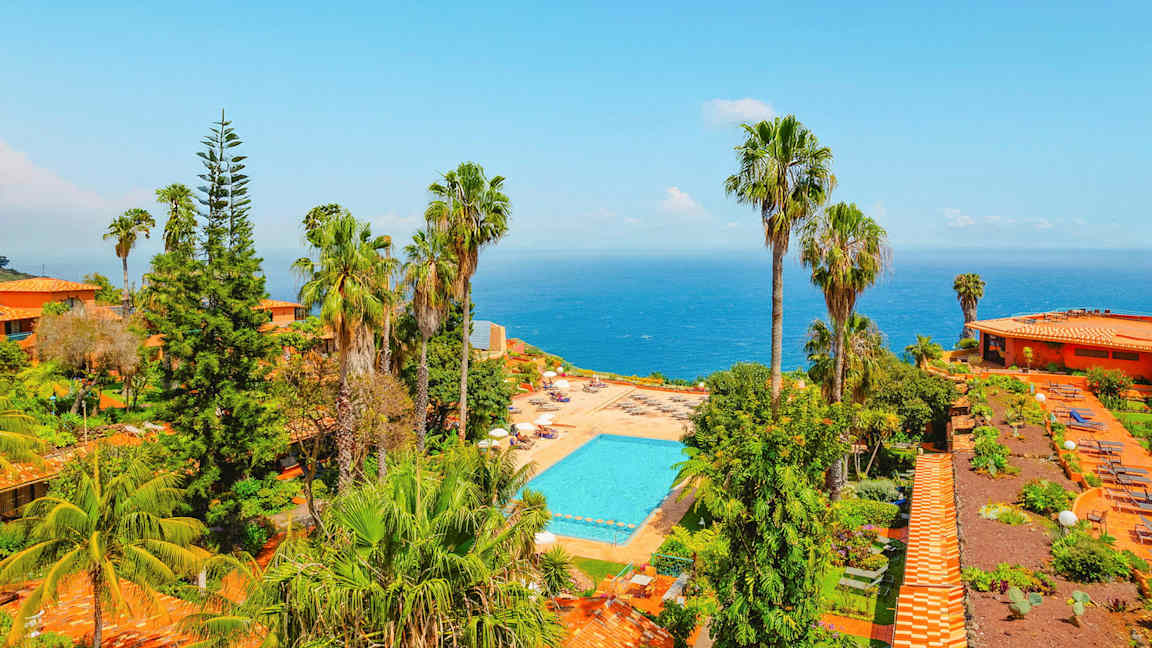 Näkymä uima-allasalueelle, jota ympäröi vehreys ja palmut, taustalla meri. (Tekoälyn luoma teksti)