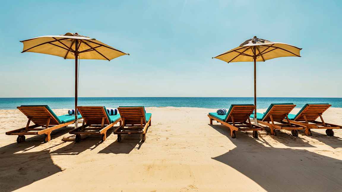 Aurinkotuoleja ja aurinkovarjoja hiekkarannalla, taustalla meri. (Tekoälyn luoma teksti)
