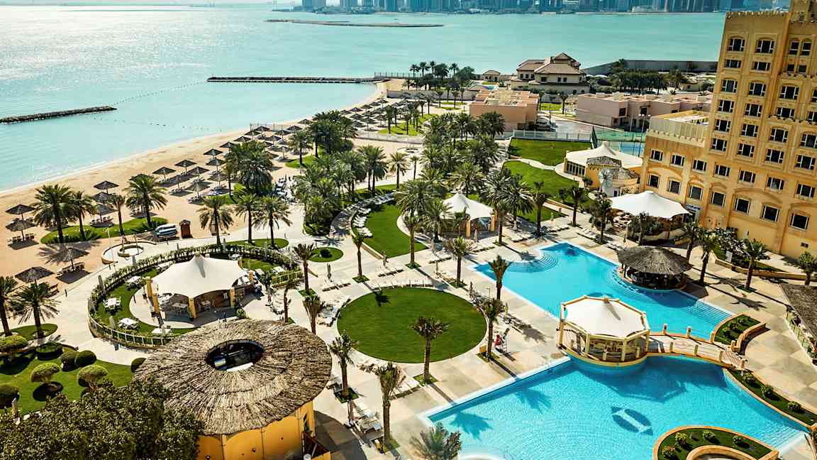 Hotellialue, jossa on uima-allas, vehreä puutarha ja näkymä rannalle sekä kaupunkinäkymiä taustalla. (Tekoälyn luoma teksti)