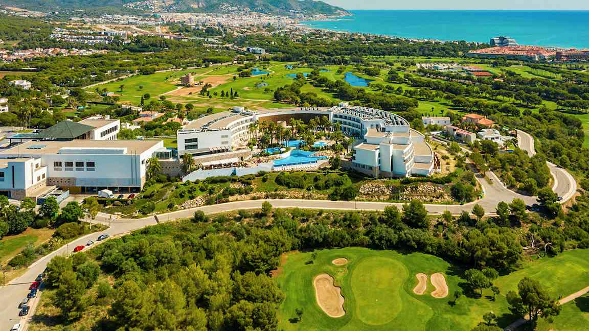 Näkymä ylhäältä hotellialueeseen, jota ympäröi vehreys ja golfkenttä. Taustalla meri ja kaupunki. (Tekoälyn luoma teksti)