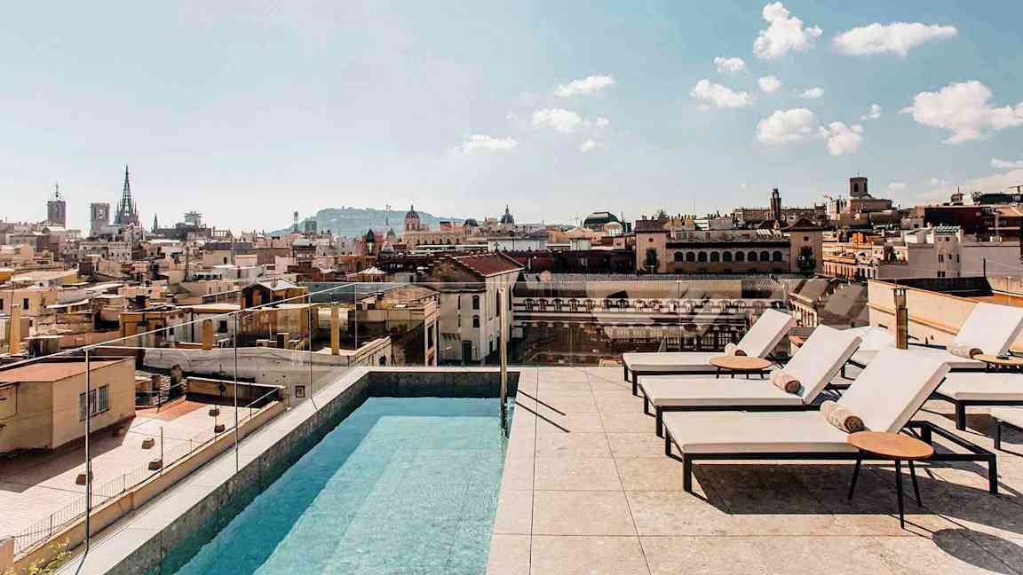 Ulkoallas ja aurinkotuoleja kattoterassilla, josta on näkymä yli kaupungin rakennusten, kirkkaalla taivaalla. (Tekoälyn luoma teksti)