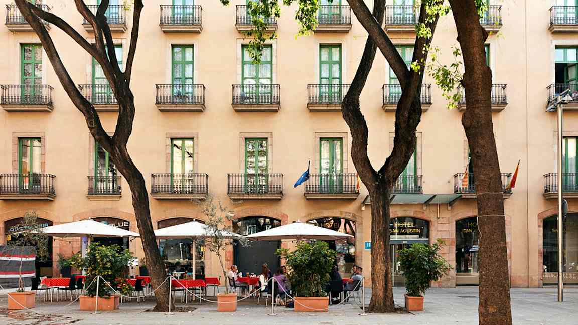 Hotelli, jossa on useita kerroksia ja parvekkeita, jalkakäytävä, jolla on aurinkovarjoja ja istumapaikkoja puiden joukossa. (Tekoälyn luoma teksti)