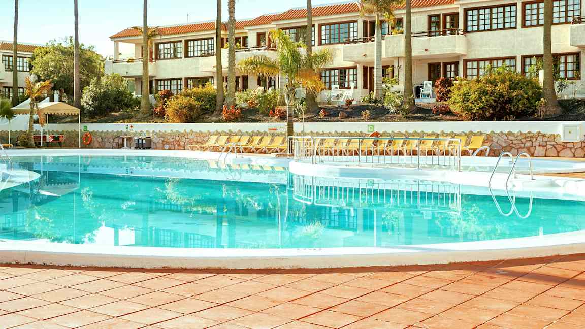 Uima-allas, jonka ympärillä on aurinkotuoleja, taustalla valkoinen rakennus. (Tekoälyn luoma teksti)