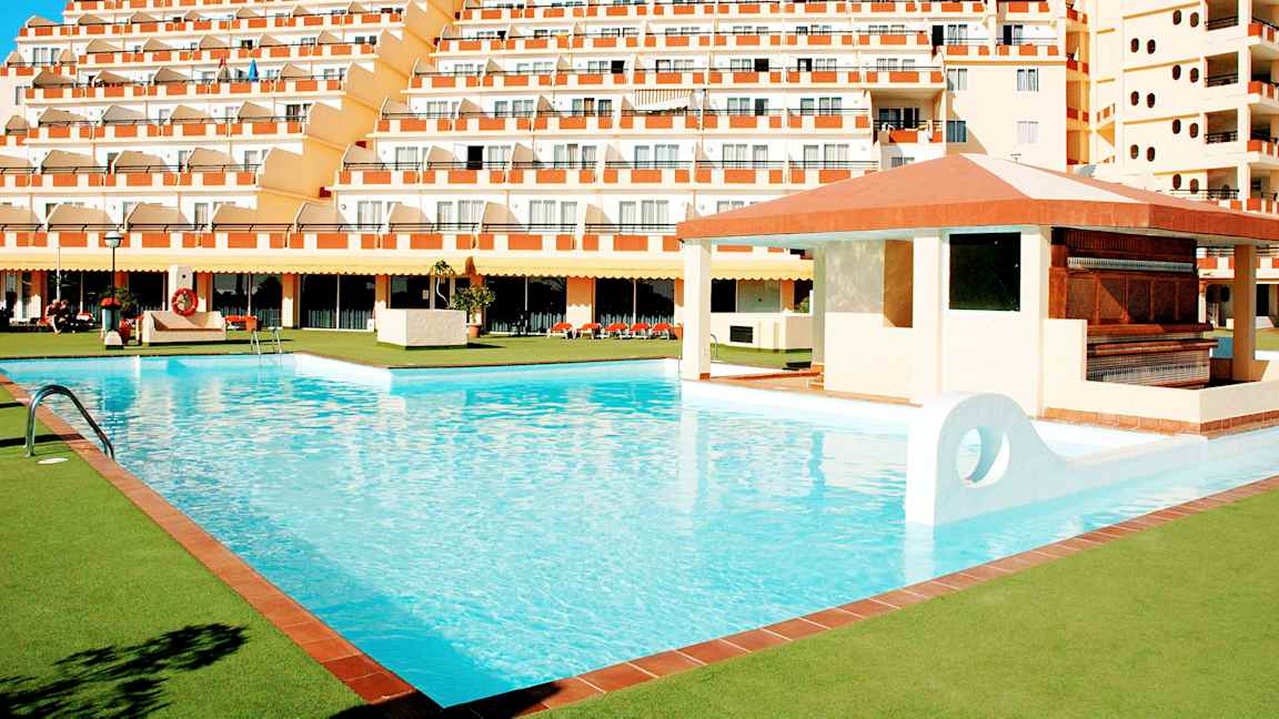 Suuri uima-allas, jota ympäröivät vihreät alueet, ja taustalla usean kerroksen hotelli. (Tekoälyn luoma teksti)