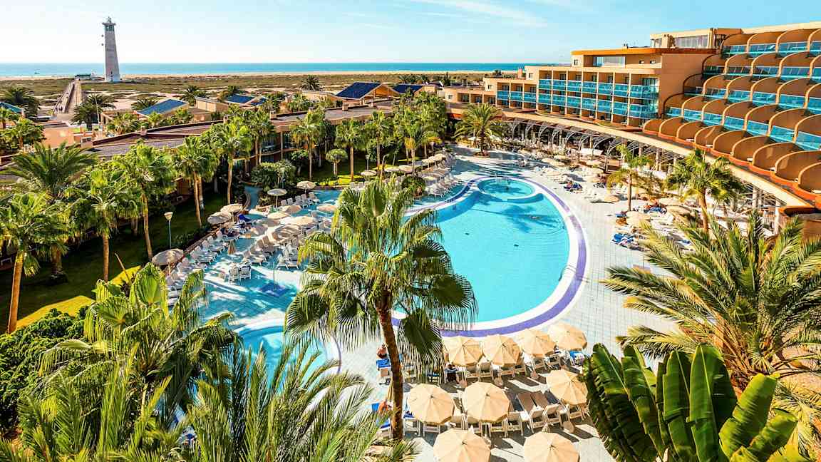 Hotellialue, jossa suuri uima-allas, aurinkotuoleja ja päivänvarjoja palmujen ympäröimänä, taustalla meri ja majakka. (Tekoälyn luoma teksti)