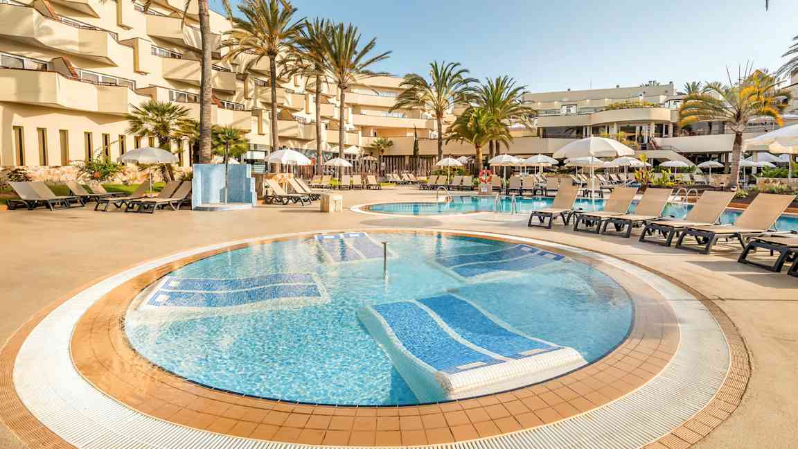 Hotellin uima-allasalue, jossa aurinkotuoleja ja aurinkovarjoja altaan ympärillä, palmupuiden ja parvekkeiden ympäröimänä. (Tekoälyn luoma teksti)