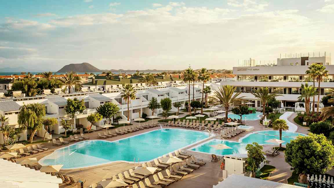 Kuva hotellialueesta, jossa on uima-altaita, aurinkotuoleja ja ympäröiviä rakennuksia sekä tulivuori taustalla. (Tekoälyn luoma teksti)