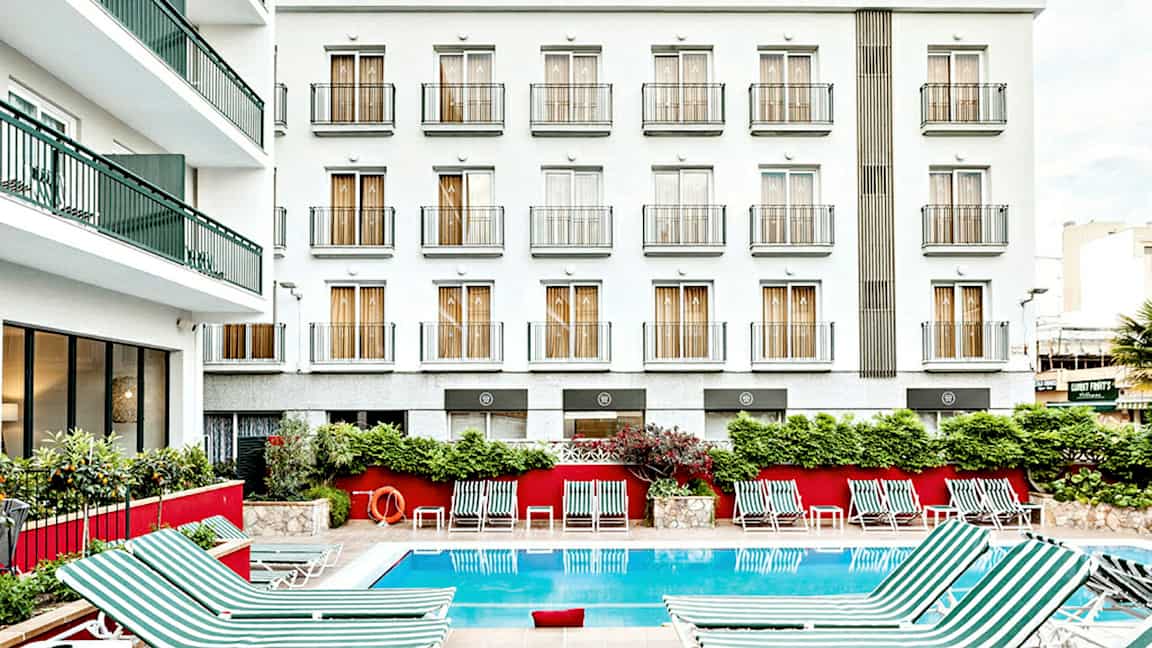 Kuva hotellirakennuksesta, jossa on valkoinen julkisivu ja useita parvekkeita. Rakennuksen edessä on ulkouima-allas, jonka ympärillä on raidallisia aurinkotuoleja. (Tekoälyn luoma teksti)