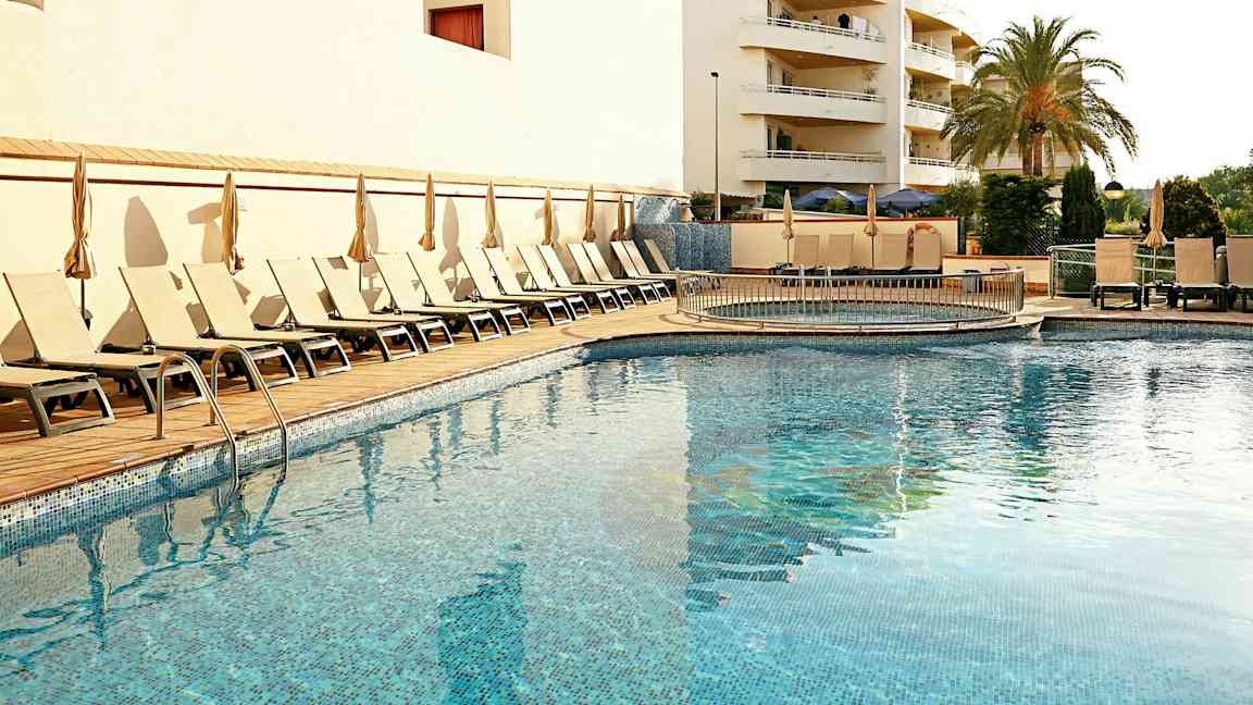 Ulkouima-allas, jota ympäröivät aurinkotuolit ja aurinkovarjot, taustalla rakennus ja palmu. (Tekoälyn luoma teksti)