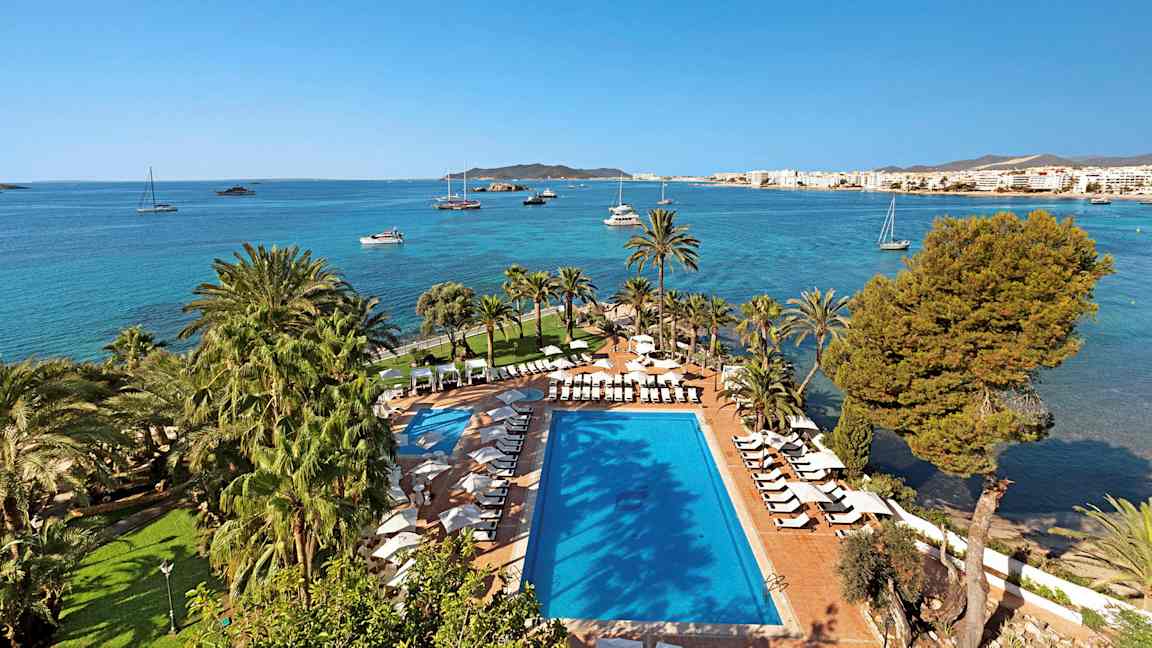 Uima-allas ja aurinkotuoleja palmujen ympäröimänä rannalla, taustalla sininen meri ja purjeveneitä. (Tekoälyn luoma teksti)