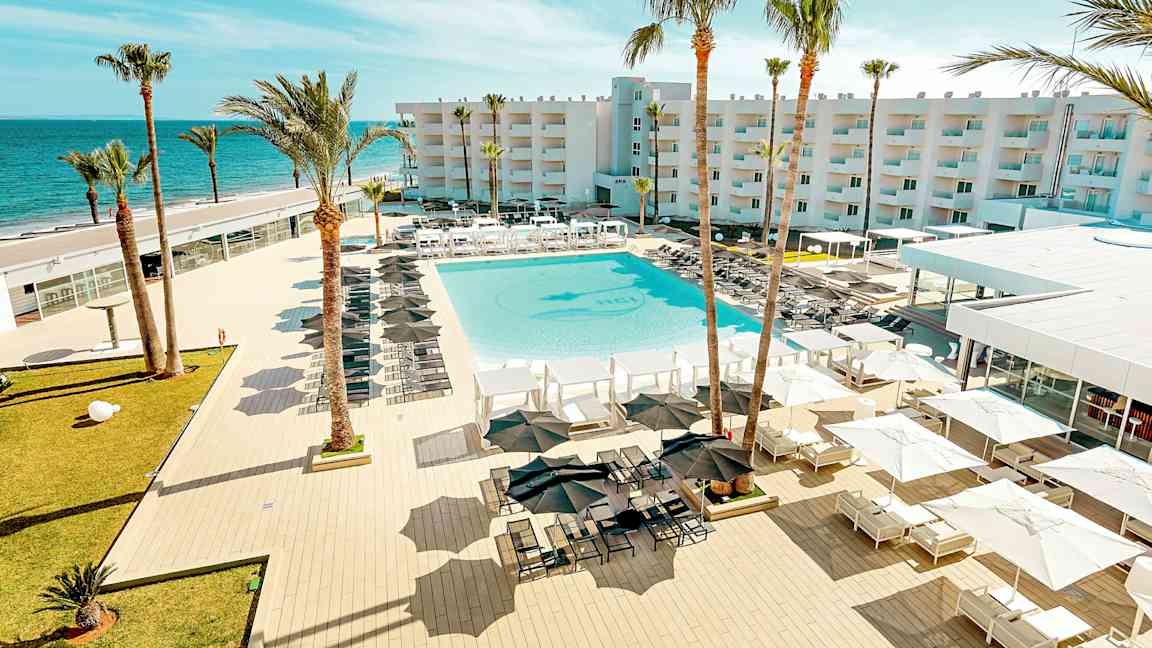 Näkymä lomakeskuksesta, jossa on suuri uima-allas, ympäröity aurinkotuoleilla ja -varjoilla, palmuja sekä rakennus taustalla lähellä merta. (Tekoälyn luoma teksti)