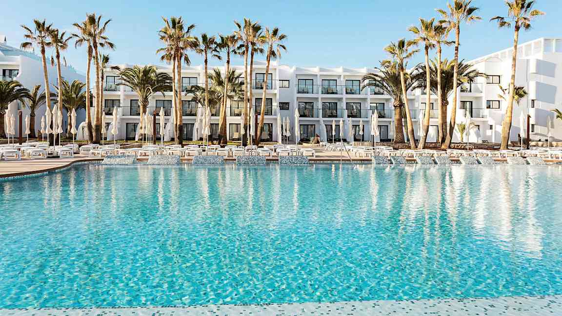 Hotellin allasalue, jossa kirkkaansinistä vettä, ympäröitynä aurinkotuoleilla, aurinkovarjoilla ja palmuilla. Taustalla näkyy valkoinen rakennus parvekkeineen. (Tekoälyn luoma teksti)