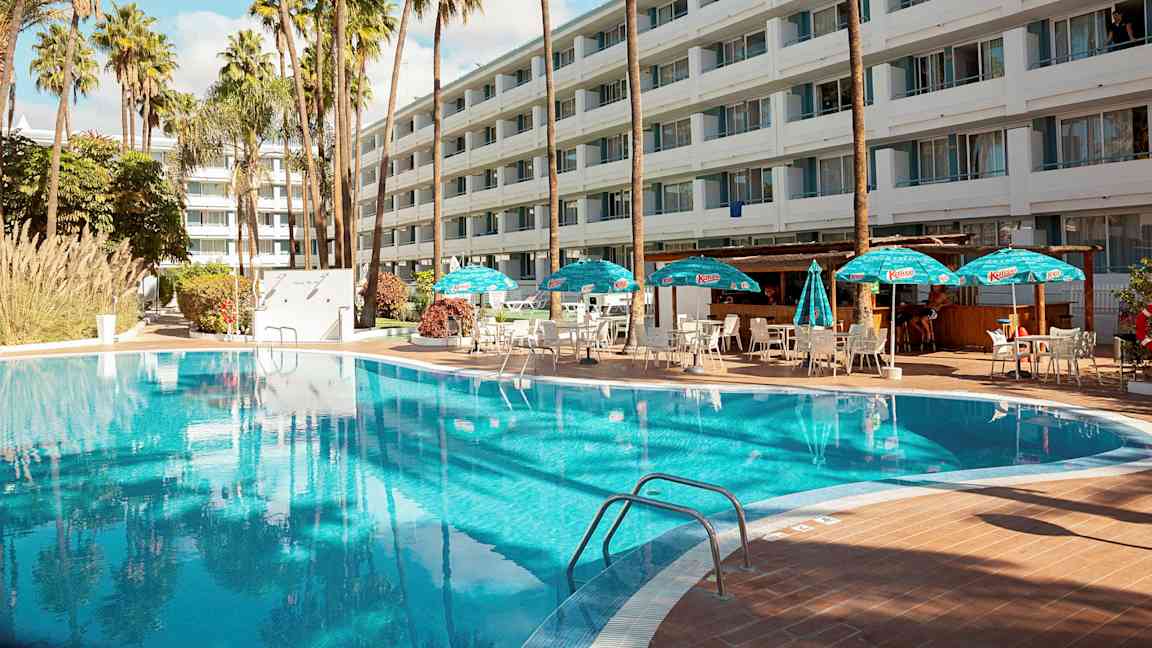 Ulkouima-allas, jossa on aurinkotuoleja ja aurinkovarjoja suuren hotellin edessä. (Tekoälyn luoma teksti)