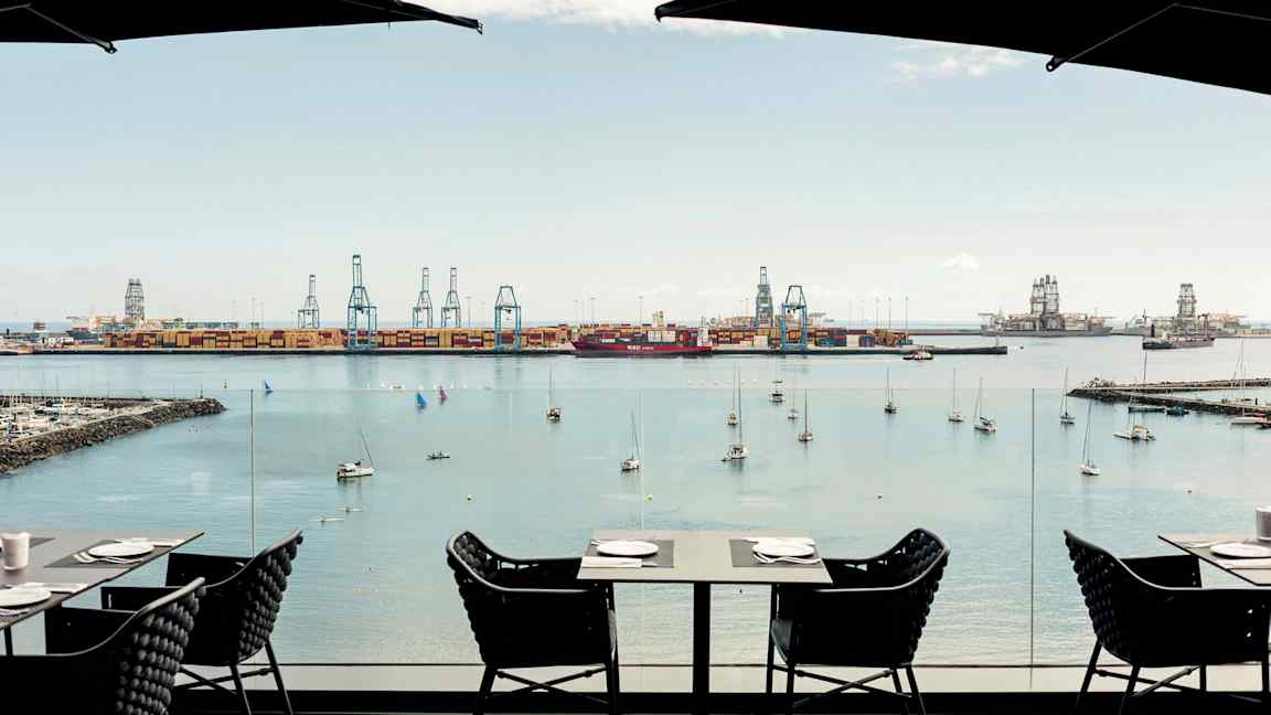 Näkymä satamaan, jossa on nostureita ja kiinnittyneitä veneitä. Etualalla on pöytiä ja tuoleja kahvilasta. (Tekoälyn luoma teksti)