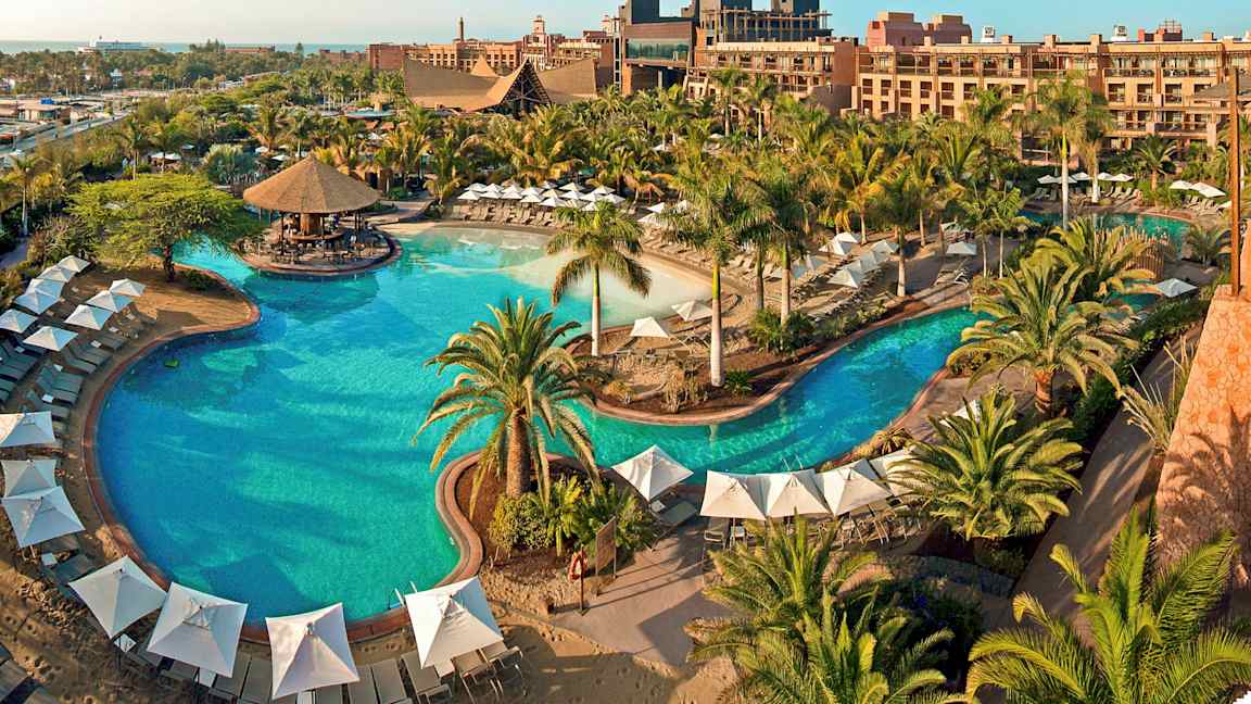 Suuri ulkoallas, jonka ympärillä on varjoja, aurinkotuoleja ja vehreää kasvustoa lomahotellissa. (Tekoälyn luoma teksti)