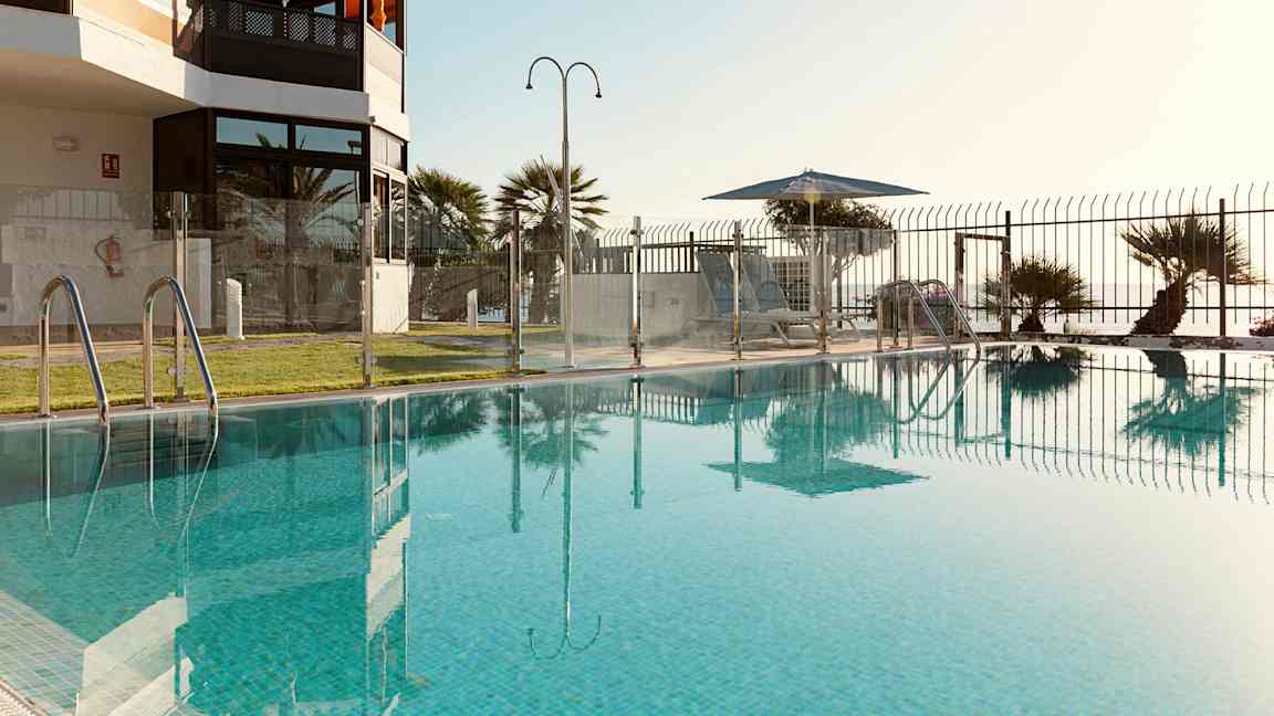 Uima-allas, jota ympäröi lasi-aita, näkymä nurmikolle ja aurinkotuoleille varjon alla. (Tekoälyn luoma teksti)