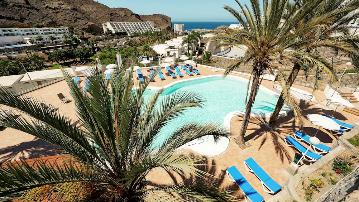 Näkymä hotellin uima-altaalle, jota ympäröivät aurinkotuolit ja palmupuut, taustalla vuoret ja meri. (Tekoälyn luoma teksti)