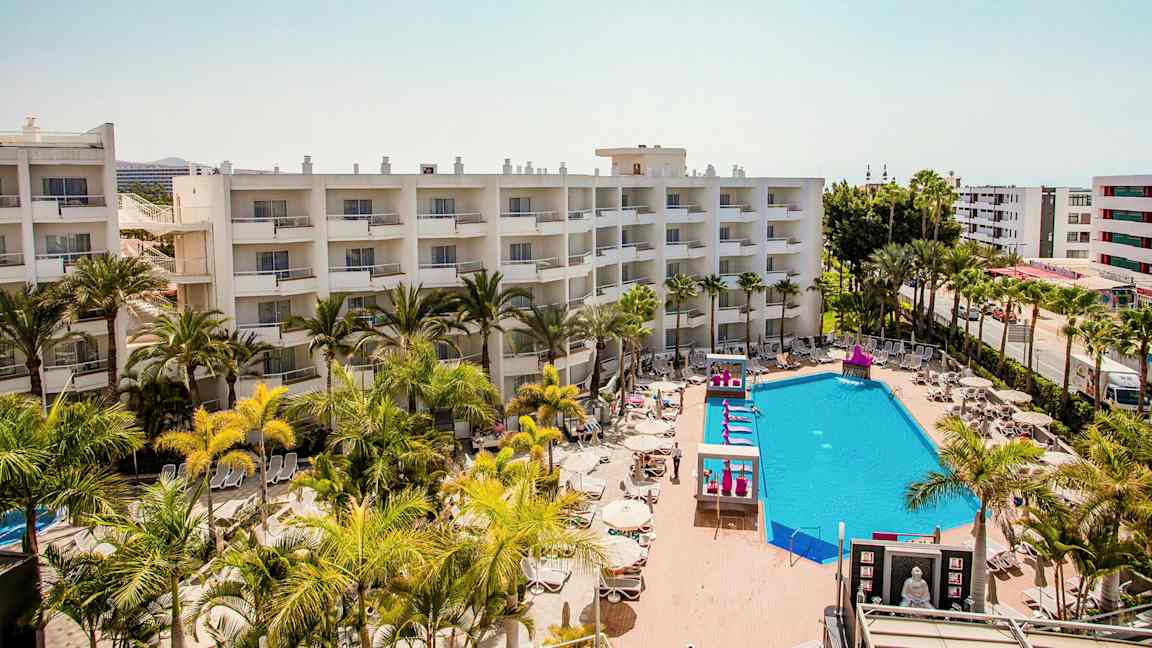 Hotellirakennukset parvekkeineen, uima-allasalue aurinkotuoleineen ja varjon alle palmuineen. (Tekoälyn luoma teksti)