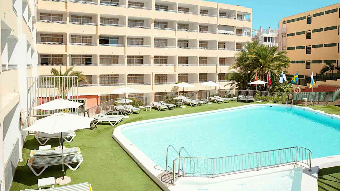 Hotellirakennus, jonka ulkopuolella on uima-allas ja aurinkotuoleja. (Tekoälyn luoma teksti)