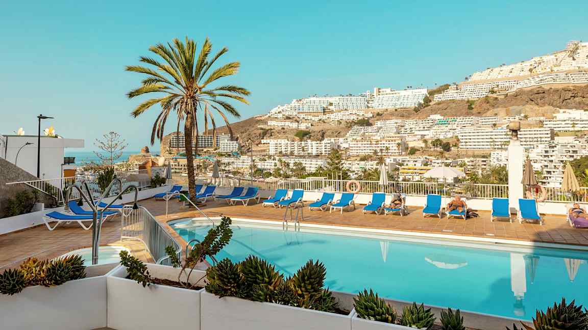 Ulkouima-allas, aurinkotuoleja ja palmuja, näkymä vuorille ja rakennuksiin. (Tekoälyn luoma teksti)