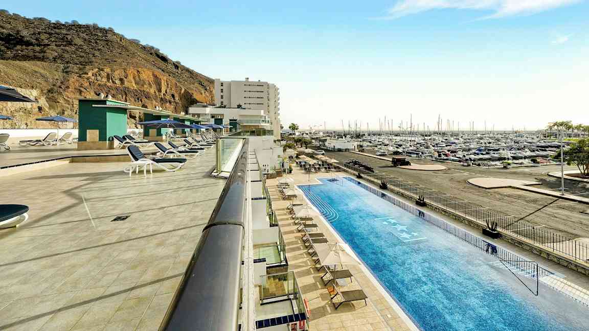 Uima-allasalue aurinkotuoleineen venesataman ja vuoren rinteen vieressä. (Tekoälyn luoma teksti)