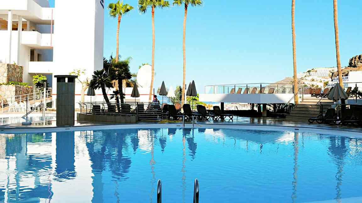 Uima-allas, jota ympäröivät aurinkotuolit, aurinkovarjot ja palmupuut, taustalla rakennus ja kirkas sininen taivas. (Tekoälyn luoma teksti)