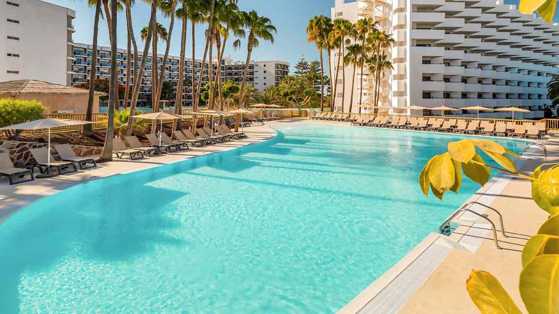 Suuri uima-allas, jota ympäröivät aurinkotuolit, aurinkovarjot ja palmut hotellin vieressä. (Tekoälyn luoma teksti)