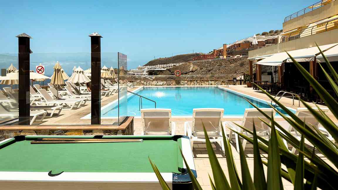 Ulkoallas, jossa on ympärillä aurinkotuoleja ja aurinkovarjoja, taustalla rakennuksia rinteessä. (Tekoälyn luoma teksti)