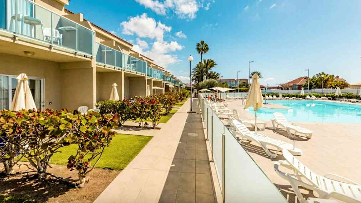 Kuva hotellialueesta, jossa on uima-allas, aurinkotuoleja ja vehreyttä. Rakennuksessa on parvekkeita. (Tekoälyn luoma teksti)