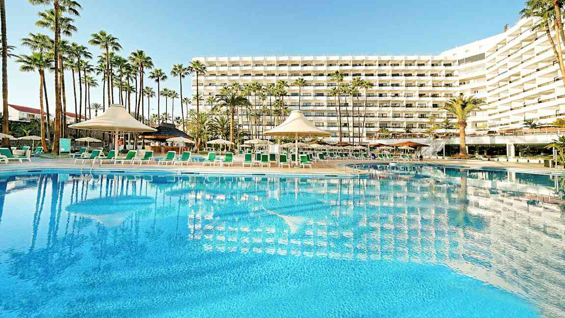 Hotellirakennus, jossa suuri uima-allas ja aurinkotuoleja etualalla, ympäröitynä palmuilla. (Tekoälyn luoma teksti)