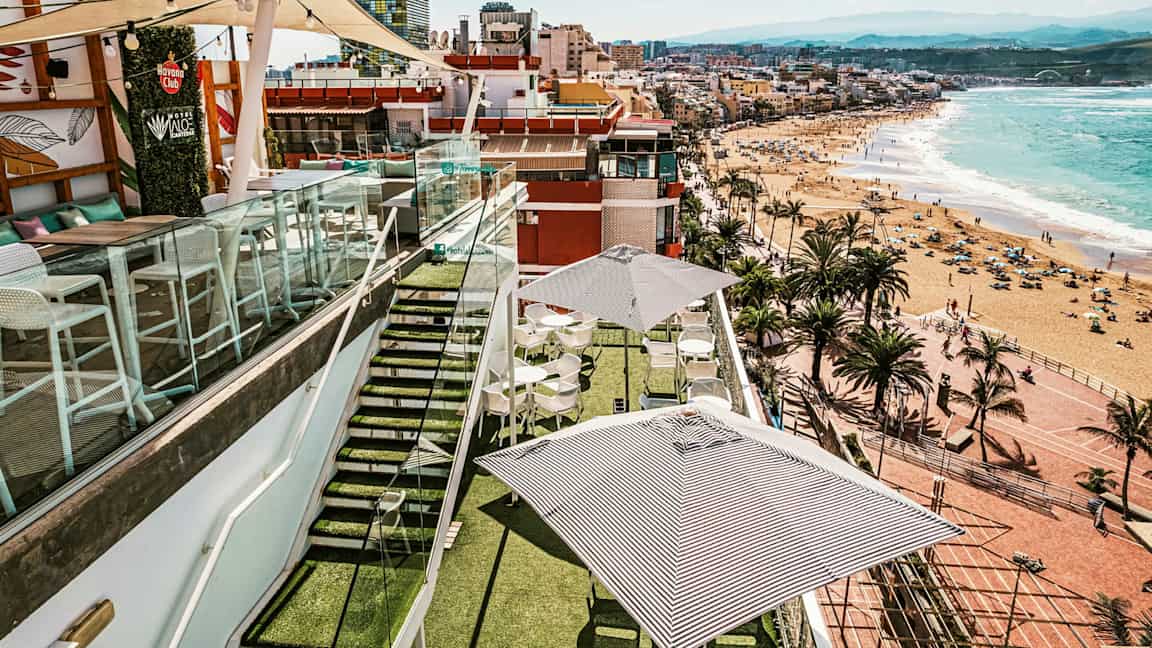 Kattoterassi, jossa pöytiä ja aurinkovarjoja, lasikaiteiden ympäröimä, näkymä rannalle ja kaupungille meren äärellä. (Tekoälyn luoma teksti)