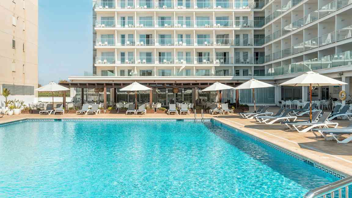 Rakennus, jossa on useita parvekkeita, uimallas edessä aurinkotuoleineen ja aurinkovarjoineen. (Tekoälyn luoma teksti)