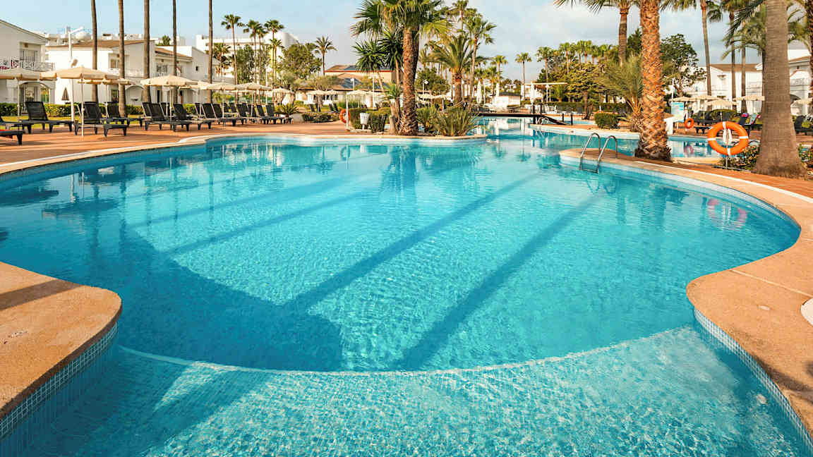 Iso ulkouima-allas, jota ympäröivät aurinkotuolit, aurinkovarjot, palmut ja rakennukset. (Tekoälyn luoma teksti)