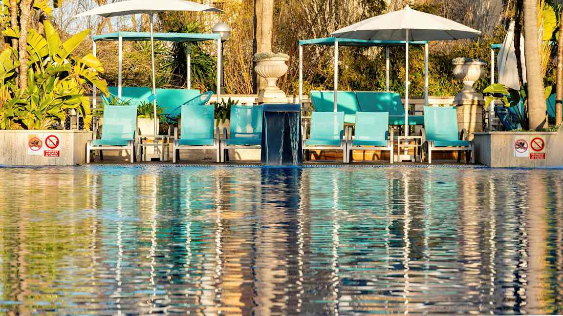 Aurinkotuolit uima-altaan äärellä, taustalla aurinkovarjoja ja vihreitä kasveja. (Tekoälyn luoma teksti)