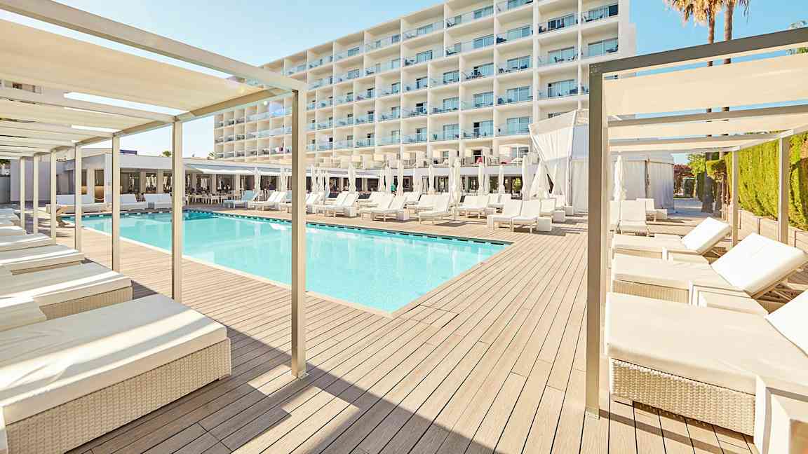 Moderni hotellikohde, jossa on uima-allas aurinkotuolien ja varjojen ympäröimänä, taustalla suuri valkoinen rakennus. (Tekoälyn luoma teksti)