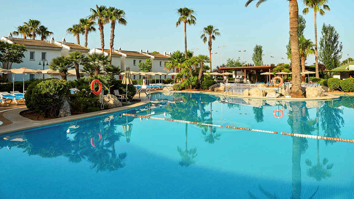 Ulkouima-allas, jota ympäröivät palmut ja aurinkotuolit, valkoiseksi maalattuja rakennuksia taustalla. (Tekoälyn luoma teksti)