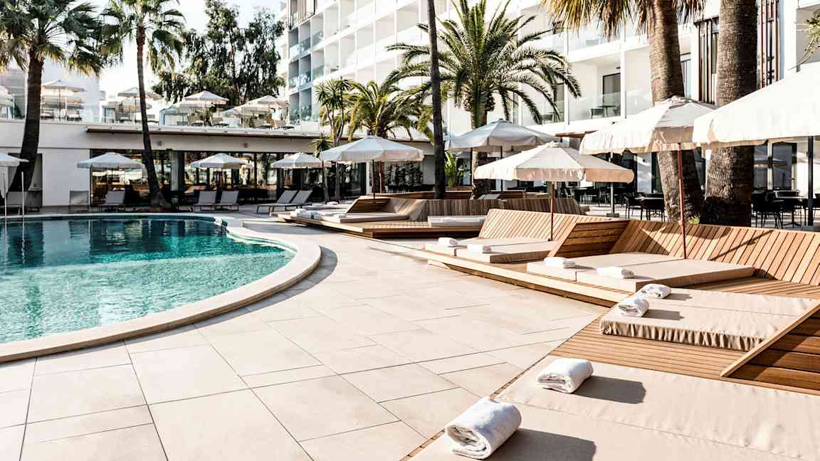 Uima-allas, jota ympäröivät aurinkotuolit ja aurinkovarjot hotellin luona, taustalla palmuja. (Tekoälyn luoma teksti)