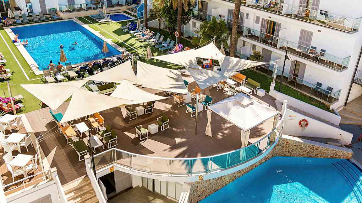 Kuva hotellilomakohteesta, jossa on suuri uima-allas aurinkotuolien ja aurinkovarjojen ympäröimänä. Etualalla on ulkobaari, jossa on paljon istumapaikkoja varjoisien kangaskatosten alla. (Tekoälyn luoma teksti)