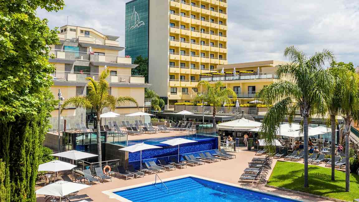 Kuva hotellista, jossa on keltaiset fasadit, ympäröivä palmuja ja uima-allas, jossa aurinkotuoleja aurinkoterassilla. (Tekoälyn luoma teksti)