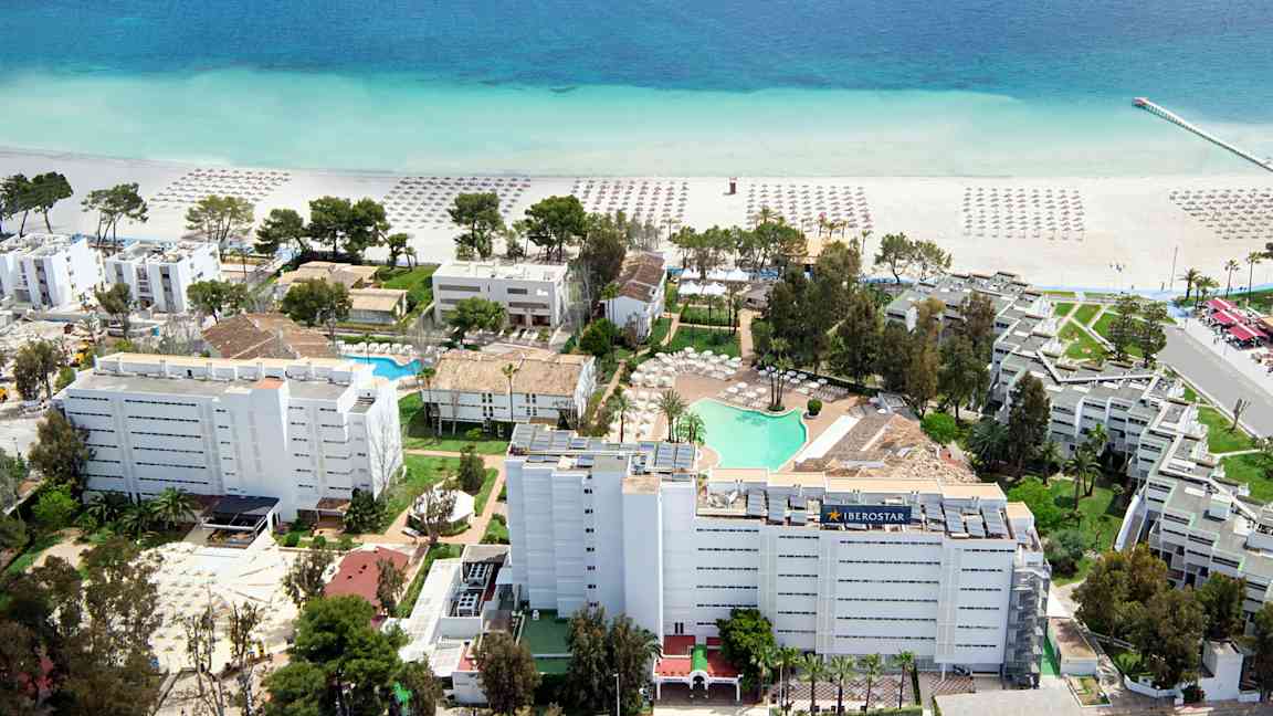 Ilmakuva hotellialueesta lähellä rantaa, jossa hiekan päällä on aurinkovarjoja ja kirkkaan sininen merimaisema. (Tekoälyn luoma teksti)