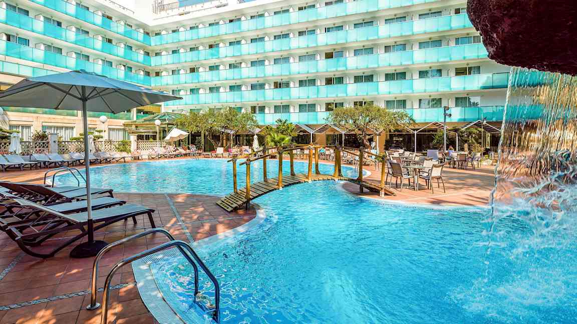 Ulkoallas hotellirakennuksen vieressä, jossa on aurinkotuoleja, pöytiä ja aurinkovarjoja. Pienen sillan yli kulkee osa vesiputousrakennelmaa. (Tekoälyn luoma teksti)