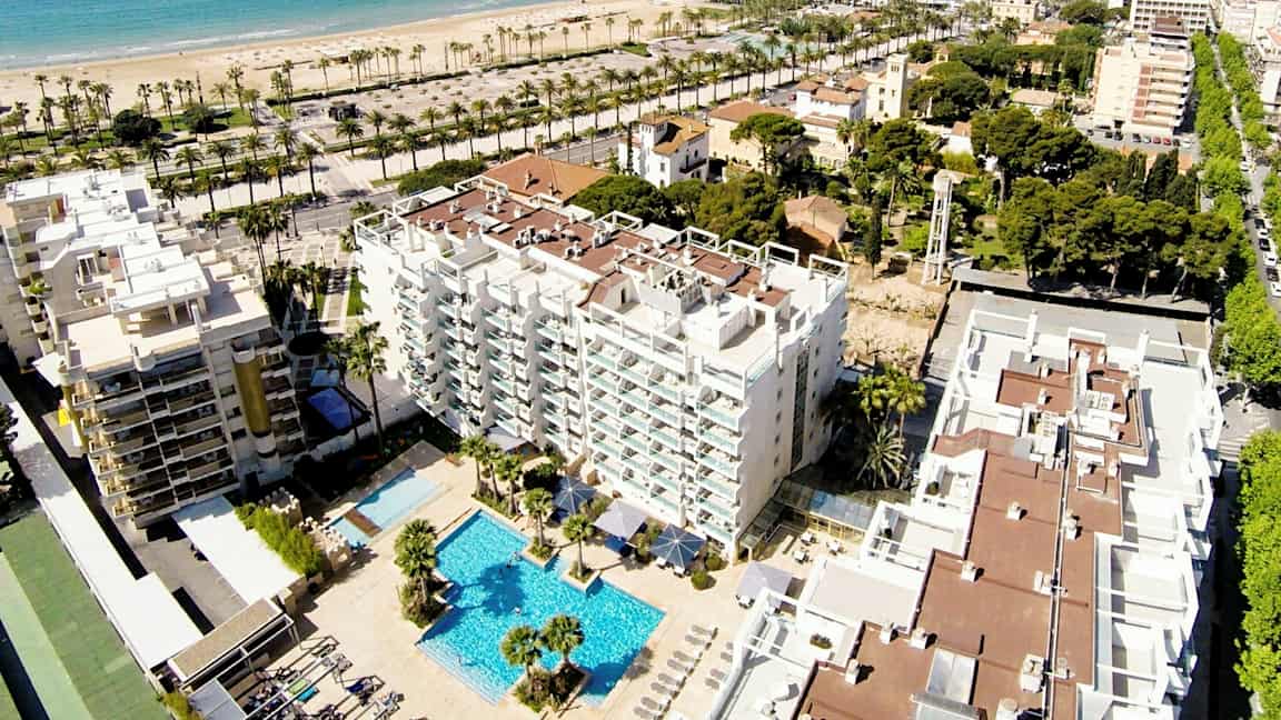 Ilmakuva hotellista, jossa on uima-allasalue, rakennusten ja vehreyden ympäröimänä. Taustalla näkyy hiekkaranta ja meri. (Tekoälyn luoma teksti)