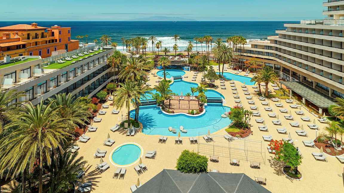 Hotellikompleksi, jossa on uima-altaita, aurinkotuoleja ja palmuja, lähellä merta. (Tekoälyn luoma teksti)