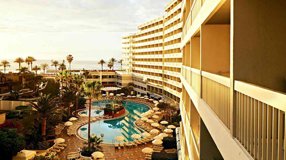 Näköala hotellin uima-altaalle, jonka ympärillä on aurinkotuoleja ja -varjoja, palmupuita sivuilla ja meri taustalla. (Tekoälyn luoma teksti)